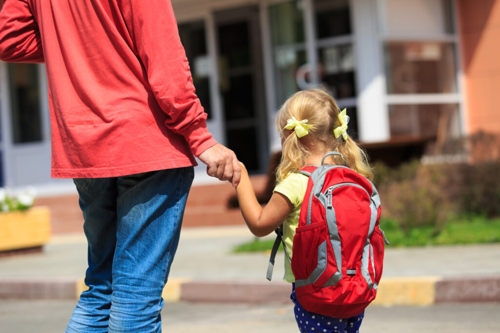 Girl holding parents hand facing away from camera, wearing backpack
