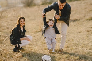 Father helping child kick soccer ball while mother watches
