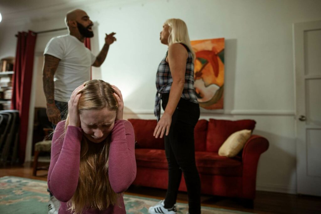 A tense family confrontation at home, featuring a distressed young girl covering her ears on the floor while an arguing couple stands in the background, highlighting the impact of family violence.